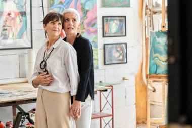 Etkilenmiş ve şık olgun bir kadın lezbiyen partneri kucaklıyor ve modern sanat atölyesine bakıyor.