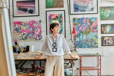 Düşünceli olgun kadın ressam renkli resimlerle sanat stüdyosunda dikilip başka tarafa bakıyor.