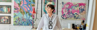 Düşünceli olgun kadın sanatçı renkli resimler ve afişlerle sanat stüdyosunda dikiliyor ve diğer tarafa bakıyor.