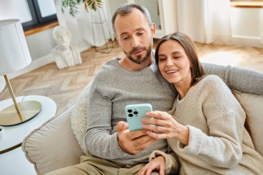Neşeli çocuksuz çift evdeki kanepede dinleniyor ve internette akıllı telefonlarda geziniyor.