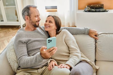 Akıllı telefonlu gülümseyen çocuksuz çift evde kanepede dinleniyor ve birbirlerine bakıyorlar.