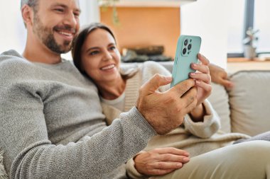 Neşeli çocuksuz çift internette akıllı telefonlarıyla geziniyor ve evdeki kanepede dinleniyorlar.
