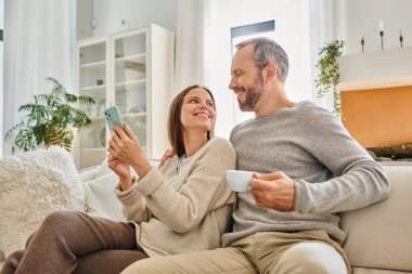 Kahve fincanı ve akıllı telefonlu mutlu çocuksuz çift oturma odasındaki kanepede birbirlerine bakıyorlar.