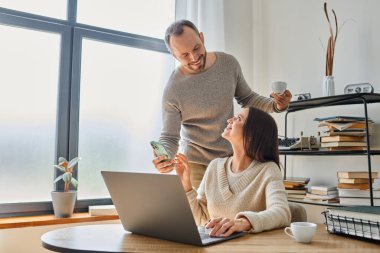 Elinde kahve fincanı olan neşeli adam ev bilgisayarında çalışan akıllı telefonu karısına gösteriyor, çocuksuz bir çift.