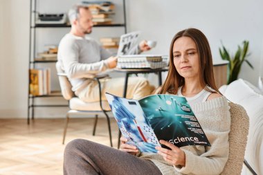 focused woman reading science magazine near husband in living room, leisure of child-free couple clipart