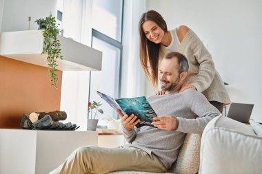joyful man reading science magazine near smiling wife on couch in living room, child-free couple clipart