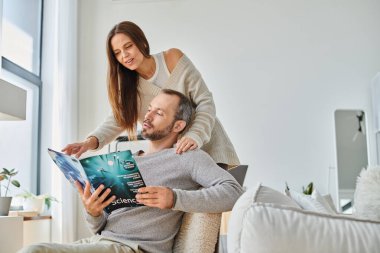 happy husband reading science magazine near smiling wife on couch in living room, child-free couple clipart