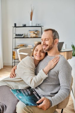 delighted woman hugging husband sitting with science magazine on couch at home, child-free life clipart