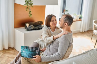 cheerful woman hugging husband sitting with science magazine on couch at home, child-free couple clipart