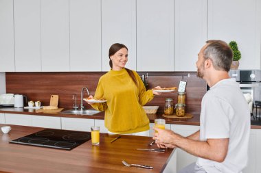 happy woman holding delicious breakfast near husband with orange juice in kitchen, child-free life clipart