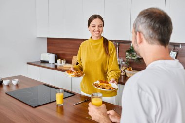 joyful wife holding delicious breakfast near husband with orange juice in kitchen, child-free couple clipart