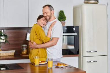 smiling child-free couple embracing near delicious breakfast in modern kitchen, love and care clipart