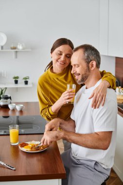 Portakal suyu içen ve gözleri kapalı bir kadın kocasını kucaklıyor mutfakta kahvaltı yapıyor, çocuksuz bir hayat sürüyor.