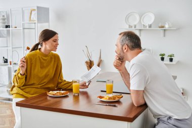 Modern mutfakta kocasıyla kahvaltı ederken gazete okuyan gülümseyen bir kadın. Çocuksuz bir çift.