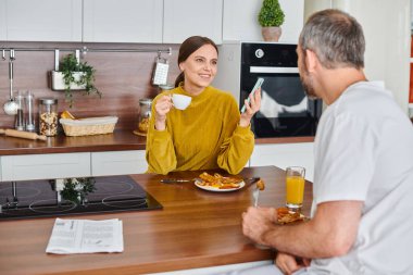 cheerful woman with coffee and smartphone talking to husband during breakfast, child-free lifestyle clipart