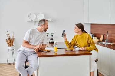 Kocasına akıllı telefon gösteren neşeli kadın modern mutfakta kahvaltı yapıyor, çocuksuz bir çift.