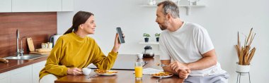 excited wife showing smartphone to husband during breakfast in modern kitchen, horizontal banner clipart