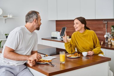 Modern mutfakta kahvaltıda kocasına akıllı telefon gösteren mutlu kadın, çocuksuz bir çift.