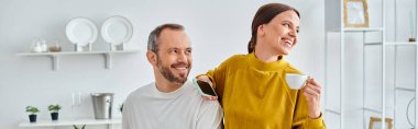 cheerful woman with morning coffee and smartphone looking away near husband in kitchen, banner clipart