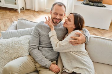 joyful woman embracing happy husband on cozy couch in living room, leisure of child-free couple clipart