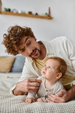 happy man with curly hair embracing his baby boy while lying together on bed, nurturing and love clipart