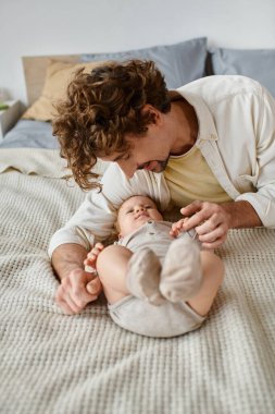 joyful father with curly hair and beard looking at his infant baby son on bed, precious moments clipart