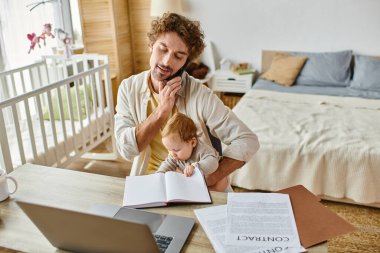 Baba küçük oğlunu kucağında tutarken akıllı telefonla konuşuyor ve evde çalışıyor, iş hayatı dengesinde.