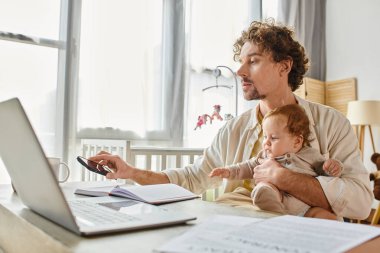 Baba, akıllı telefonunu alıp evde çalışırken bebeğini kucağına alıyor. İş hayatı dengesi.
