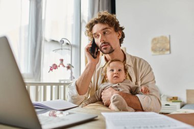 Baba küçük oğlunu kucağında tutarken akıllı telefonla konuşuyor ve evdeki kağıt ve dizüstü bilgisayarların yanında çalışıyor.