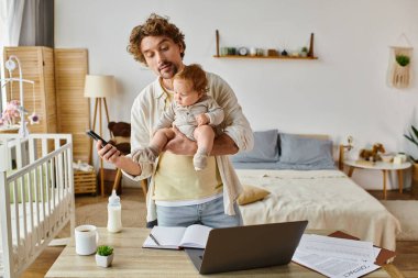 busy single dad holding infant son and using smartphone near laptop and baby bottle on desk clipart