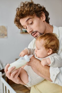 curly-haired father attentively feeding his infant son with milk in baby bottle, fatherhood and care clipart
