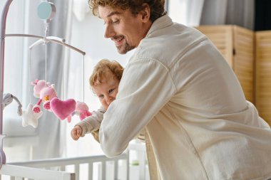 caring single father holding his infant son near baby mobile in nursery, fatherhood and love clipart