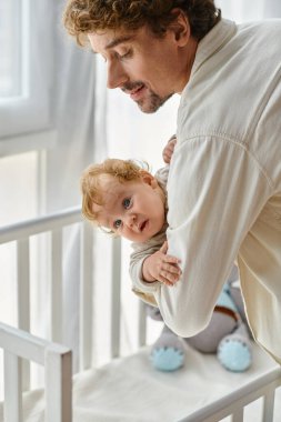 loving and happy father lifting his infant son with blue eyes from baby crib in nursery, fatherhood clipart