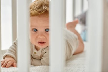 Mavi gözlü erkek bebek, bebek odasındaki döşemeleri dikizliyor, masumiyetini koruyor.