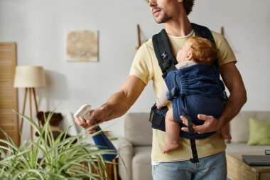 cropped shot of father with infant son in carrier holding spray bottle and watering green plant clipart