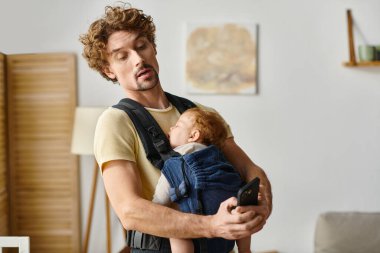 kıvırcık saçlı bekar baba akıllı telefonda mesajlaşırken bebek taşıyıcıda uyuyor, modern ebeveynlik