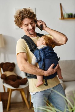 Neşeli baba akıllı telefonda konuşuyor. Bebek taşıyıcıda uyuyor. Hayat ve iş arasında denge var.