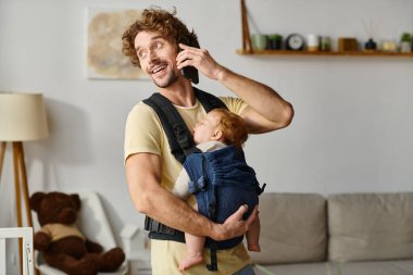 Akıllı telefondan konuşan neşeli bir adam. Bebek taşıyıcıda uyuyor. Hayatla iş arasındaki denge.