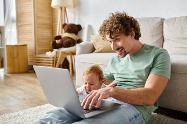 cheerful father using laptop near infant son in living room, balancing between fatherhood and job clipart