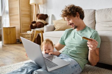 Kıvırcık saçlı mutlu bir adam, oturma odasında çocuğun yanında internetten alışveriş yaparken elinde kredi kartı tutuyor.