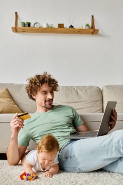 Mutlu babanın elinde kredi kartı varken çıngıraklı yılanla oynayan çocuğun yanında online alışveriş yapıyor.