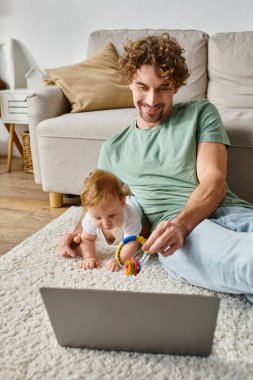 happy man holding baby rattle near infant son and laptop on carpet, balancing between work and life clipart