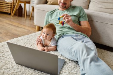 joyful man holding baby rattle near infant son and laptop on carpet, balancing between work and life clipart