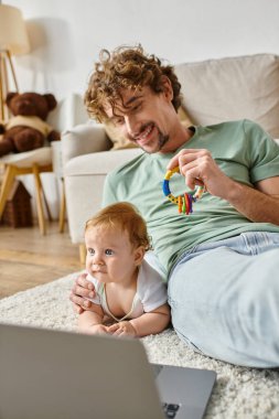 Bebek çıngırağı tutan neşeli bir adam mavi gözlü, laptoplu, iş ve yaşam dengesi olan bir çocuğun yanında.