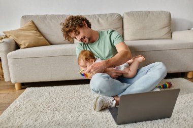 happy man with curly hair holding baby rattle near infant son and laptop, work and life balance clipart