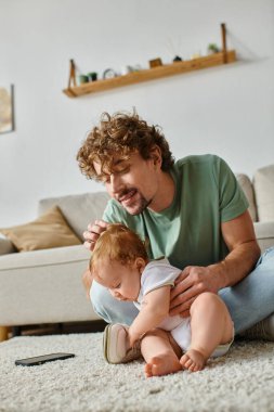 Mutlu bekar baba evde halıda bebekle otururken akıllı telefon kullanıyor.