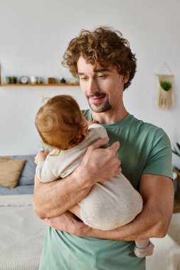 Kıvırcık saçlı ve sakallı şefkatli baba kucağında bebek kıyafetleri içindeki oğlu, ebeveynlik yapıyor.