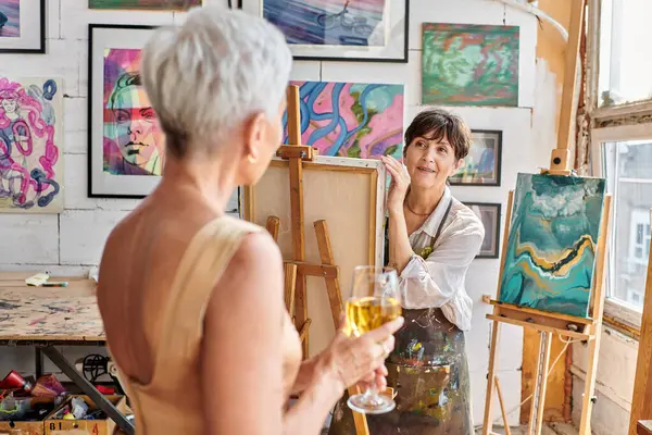 Sanat atölyesinde şarap kadehiyle poz veren bulanık orta yaşlı bir modele bakan esinlenmiş kadın sanatçı.