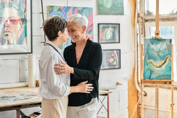 happy and fashionable lesbian artists embracing in modern art studio with creative paintings