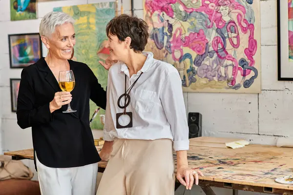 在现代艺术工作室里 一个快乐而时尚的成熟女人 带着酒杯和女同志聊天 — 图库照片